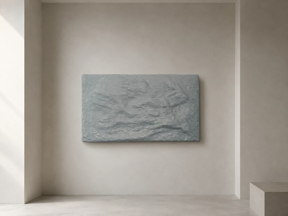 A pale grey-blue relief the shape of a still water surface, hung centred on a warm white gallery wall