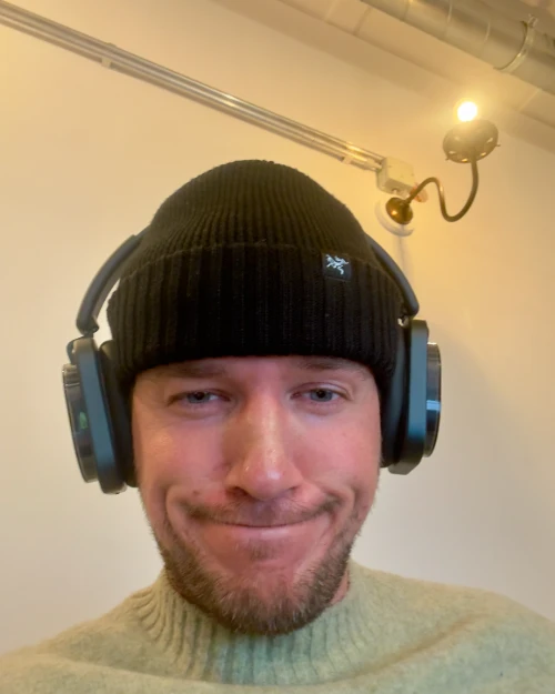 Thore at home: black beanie, over-ear headphones, light green jumper, warm white wall behind